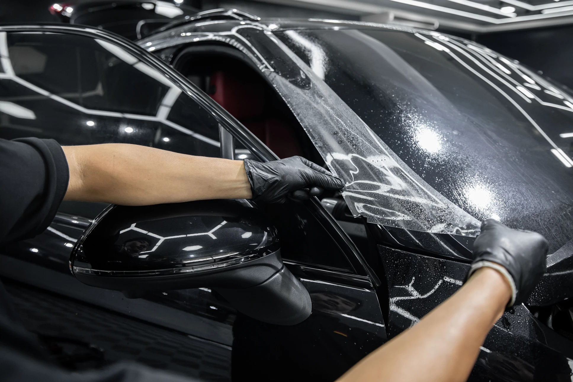 Intérieur atelier automobile, technicien appliquant film de protection transparent sur carrosserie noire brillante d'un véhicule