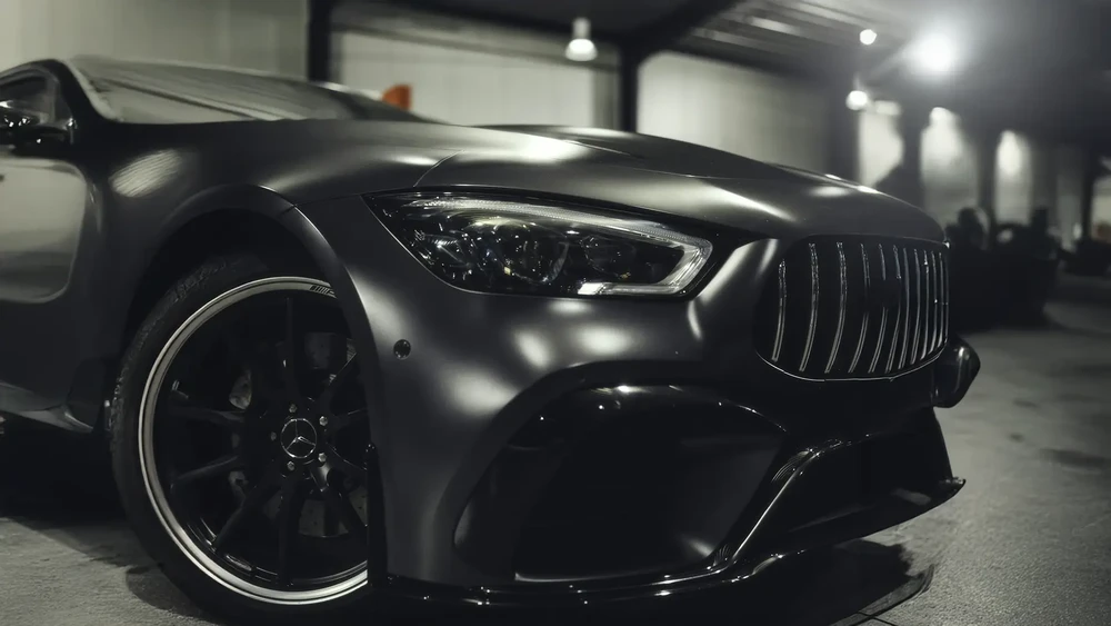 Homme en costume bleu à carreaux étreignant affectueusement le capot d'une voiture grise dans un showroom automobile