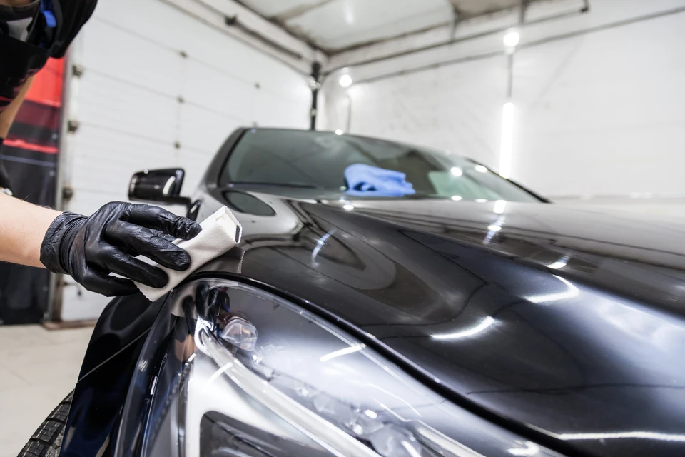Intérieur d'atelier automobile, technicien en gants nitrile noirs applique produit de protection céramique sur carrosserie noire avec applicateur microfibre