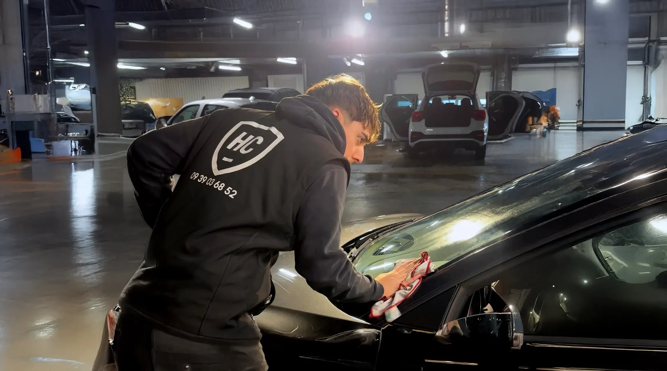 Atelier automobile intérieur avec technicien en uniforme rouge et casquette nettoyant minutieusement un véhicule sombre